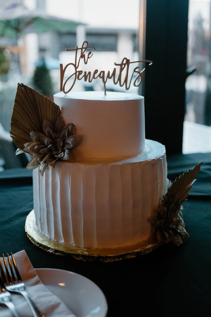 Two-tier white wedding cake with gold topper reading The Deneaults and dried botanical accents